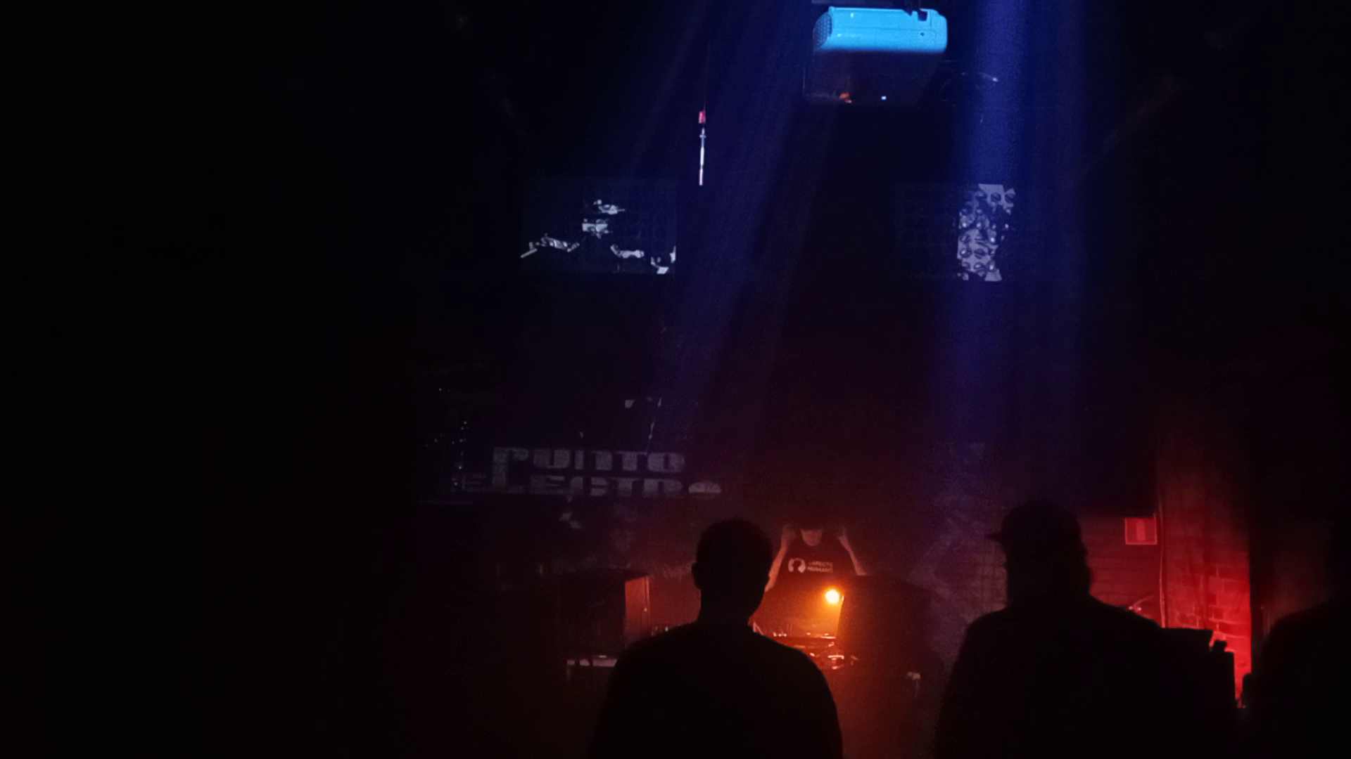 Punto Electro live set, desk light illuminating DJ setup, dimly lit room, punto electro logo visible behind and projector mounted to ceiling