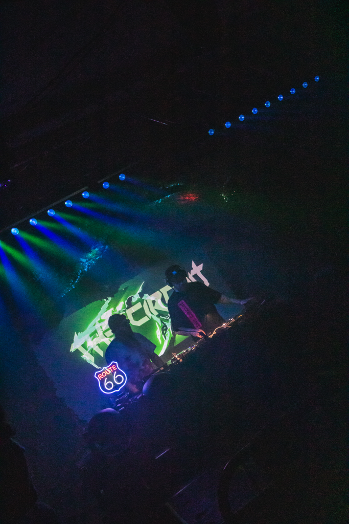 The Circuit DNB Live set 2 DJs performing DJs OG Otter and Sheidow, camera is far away and tilted angle, Circuit logo like warped energy field behind performers and Route 66 glowing light on the table